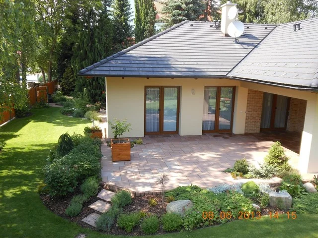 Dlážděná terasa domu IRIDA s okrasnými záhony a trávníkem