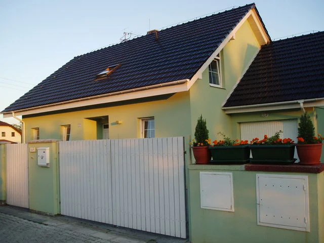 Dům APOLLON od ulice s bílou vjezdovou branou a truhlíky květin