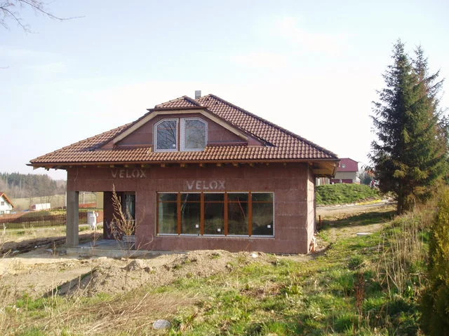 Rozestavěný dům Charis s červenou střechou a smrkem vedle stavby