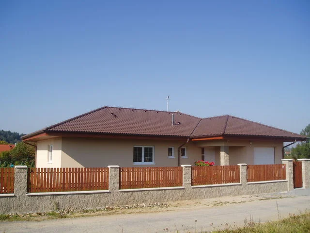 Bungalov IRIS s vínově hnědou valbovou střechou za dřevěným plotem