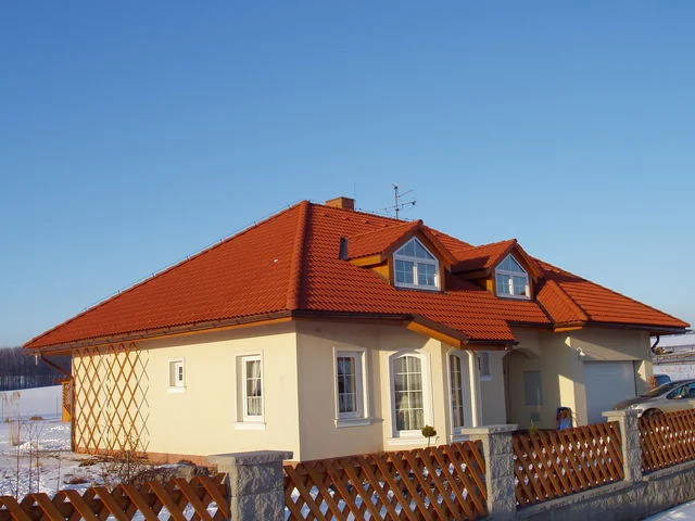 Dům AFRODITE 1 za dřevěným plotem, červená valbová střecha s vikýři v zimě