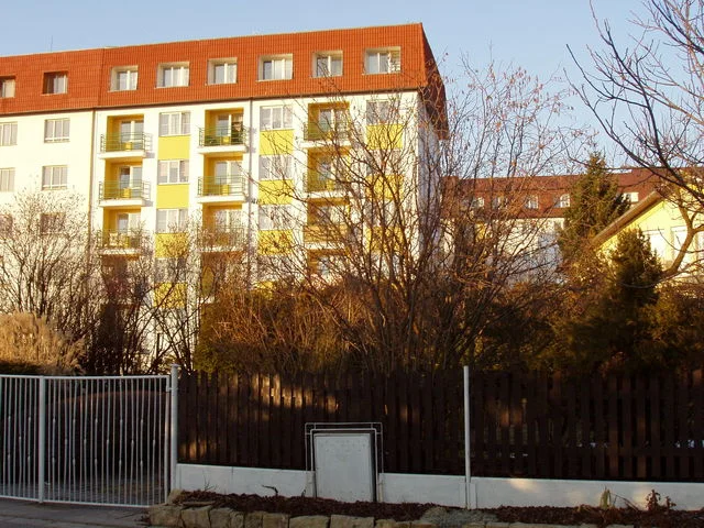 Bytový dům Větrník za dřevěným plotem a holými stromy v podvečer