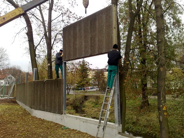 Dva pracovníci na štaflích osazují panel stěny nad vodním tokem