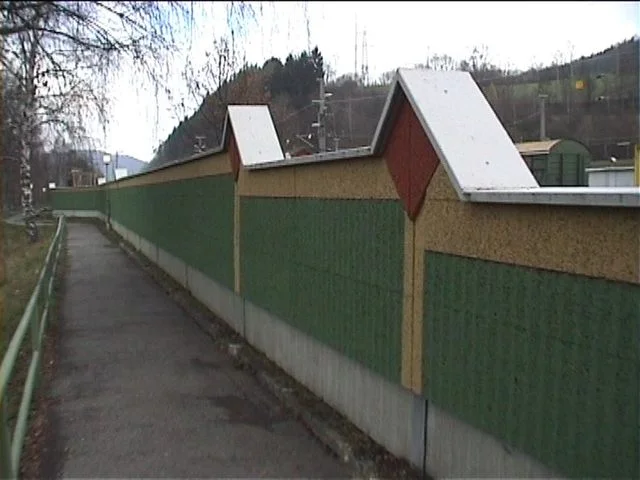 Zelená protihluková stěna s bílými sedlovými stříškami podél pěší cesty